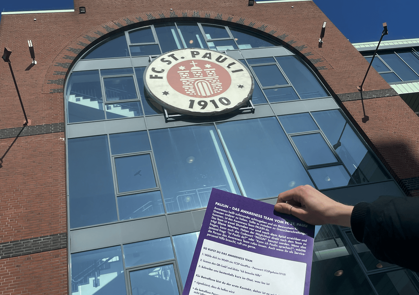 Hand hält lila Infoblatt vor dem Eingang des FC St. Pauli Stadions mit großem Vereinslogo.