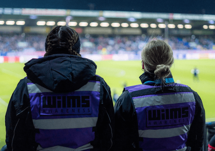 Zwei Personen von hinten gesehen, die auf einer Tribüne sitzen und auf ein Fußballspiel schauen. Sie tragen Jacken mit der Aufschrift "Wilms Awareness" in lila und schwarz. Im Hintergrund ist das beleuchtete Fußballfeld mit unscharfen Zuschauern zu erkennen.