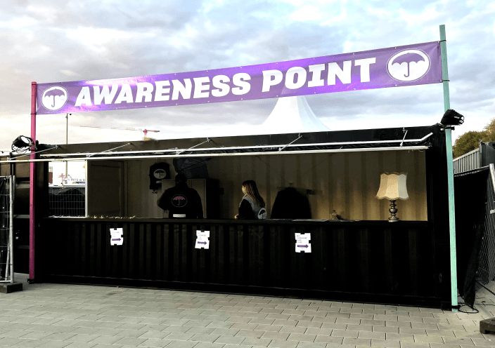 Ein Informationsstand im Freien mit der Aufschrift 'AWARENESS POINT' auf einem lila Banner. Der Stand ist mit einer schwarzen Theke ausgestattet, hinter der zwei Personen stehen. Auf der Theke befinden sich ein Laptop und eine Tischlampe. Der Stand ist beleuchtet und der Himmel im Hintergrund zeigt eine Dämmerung.